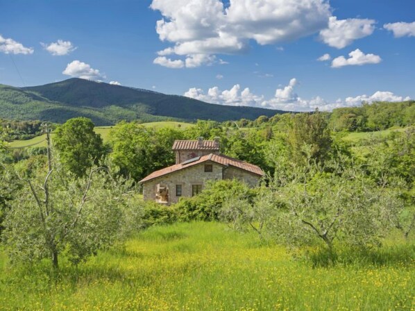 A stone house with a red roof surrounded by lush greenery and hills in the background.