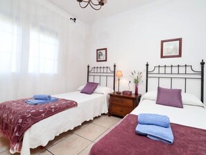 Une chambre d’hôtel avec deux lits, chacun agrémenté d’un oreiller violet et d’une couvre-lit blanche. Une table de chevet avec une lampe et un vase de fleurs se trouve entre les deux lits.