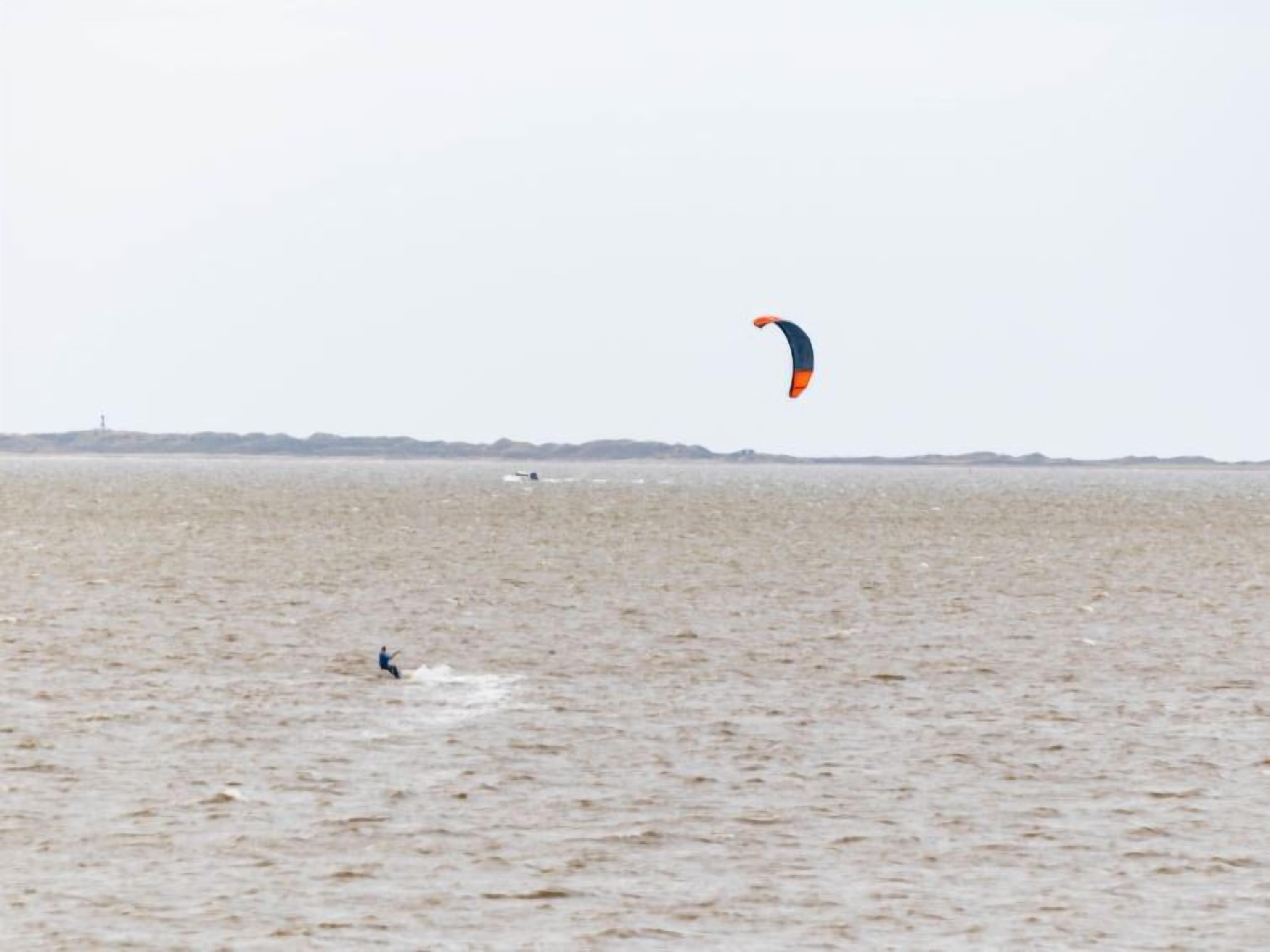 Una persona practica windsurf en una extensa masa de agua, con una costa distante y un faro.