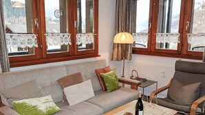 Un salon chaleureux avec un canapé, un fauteuil et une table basse. Les fenêtres sont ornées de rideaux en dentelle, et une lampe est posée sur la table.