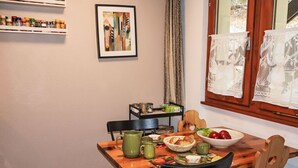 Un coin repas avec une table en bois, des chaises, un saladier de fruits et une étagère remplie d’épices.