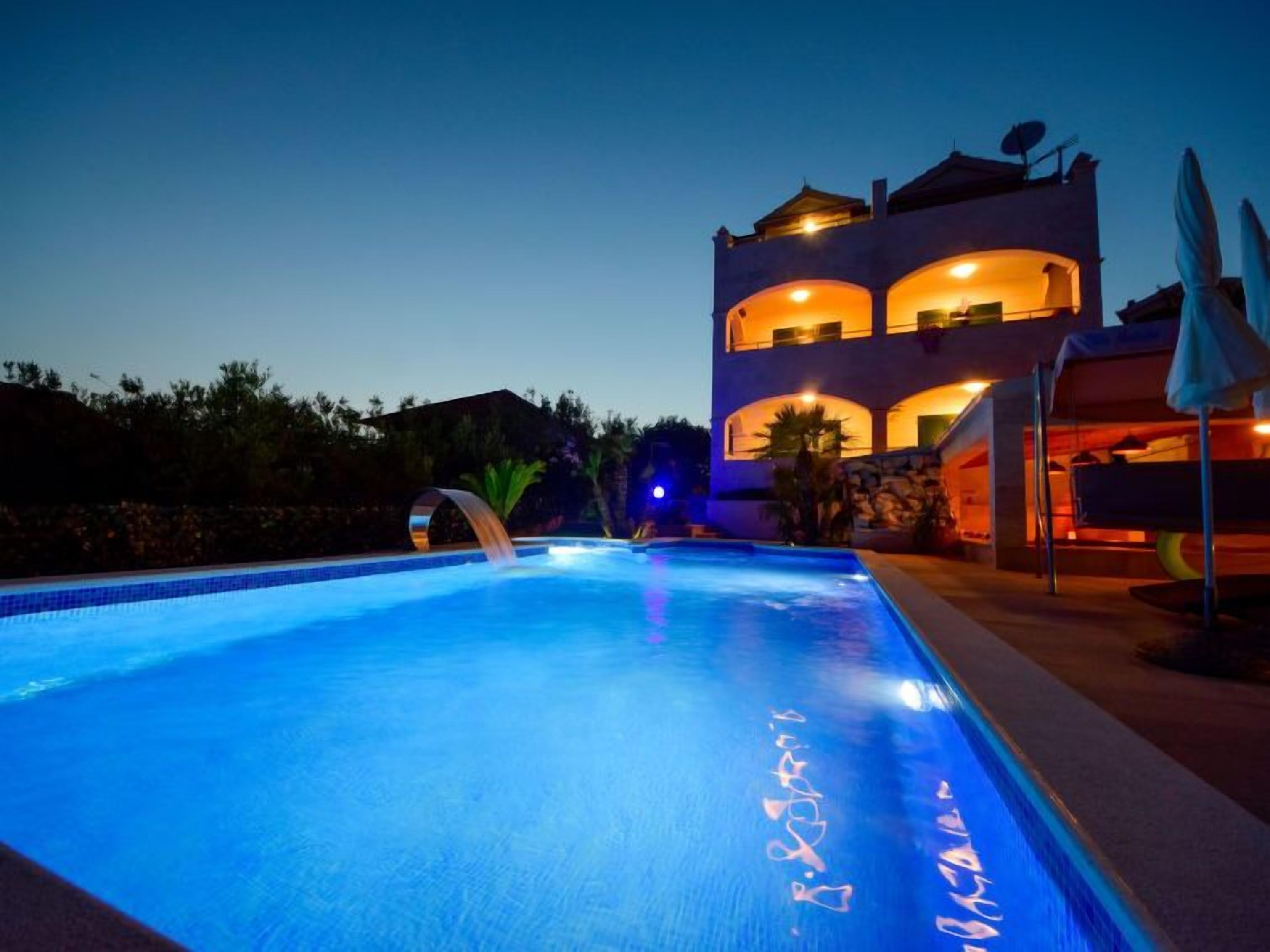 A swimming pool with a waterfall feature, surrounded by a two-story building with balconies and outdoor umbrellas.