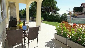 A covered patio with a table and chairs, a grill, and a garden with a wooden fence. - Apartment Ante by Interhome (Brodarica)