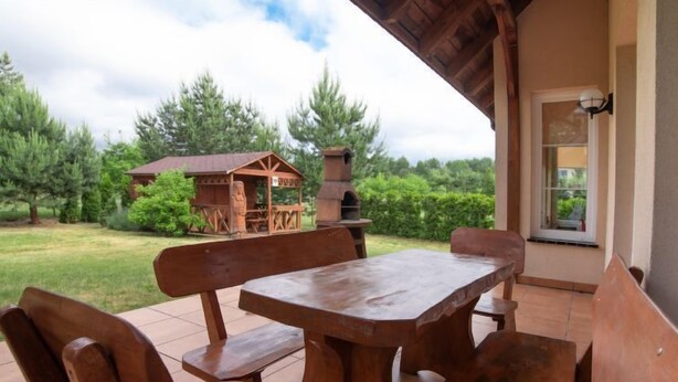 An outdoor patio with wooden benches and a table, surrounded by a garden and a wooden structure in the background.
