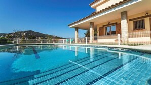 Une piscine à l’eau d’un bleu limpide, entourée d’une clôture blanche. Au fond, on aperçoit un bâtiment avec un toit en tuiles et une vue sur une colline parsemée de maisons.