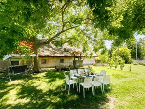 A rustic stone house with a thatched roof, surrounded by a lush garden and a dining table set for six with white chairs.