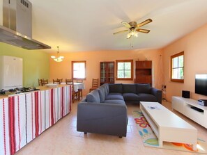 A modern living room with a sofa, a dining area with a table and chairs, a kitchen with a stove and cabinets, and a ceiling fan.