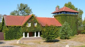 Une maison avec un toit à forte pente recouvert de lierre, une tour imposante et un chemin de pierre menant à l’entrée.