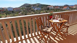 Un balcon avec une table et des chaises en bois, un vase rouge rempli de fleurs et une vue sur une colline parsemée de maisons.