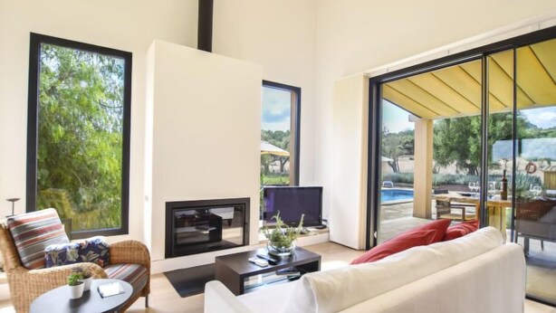 Una sala moderna con un sofá, un sillón, una mesa de centro y un televisor. Hay un ventanal y un portón de cristal que dan a un espacio exterior.