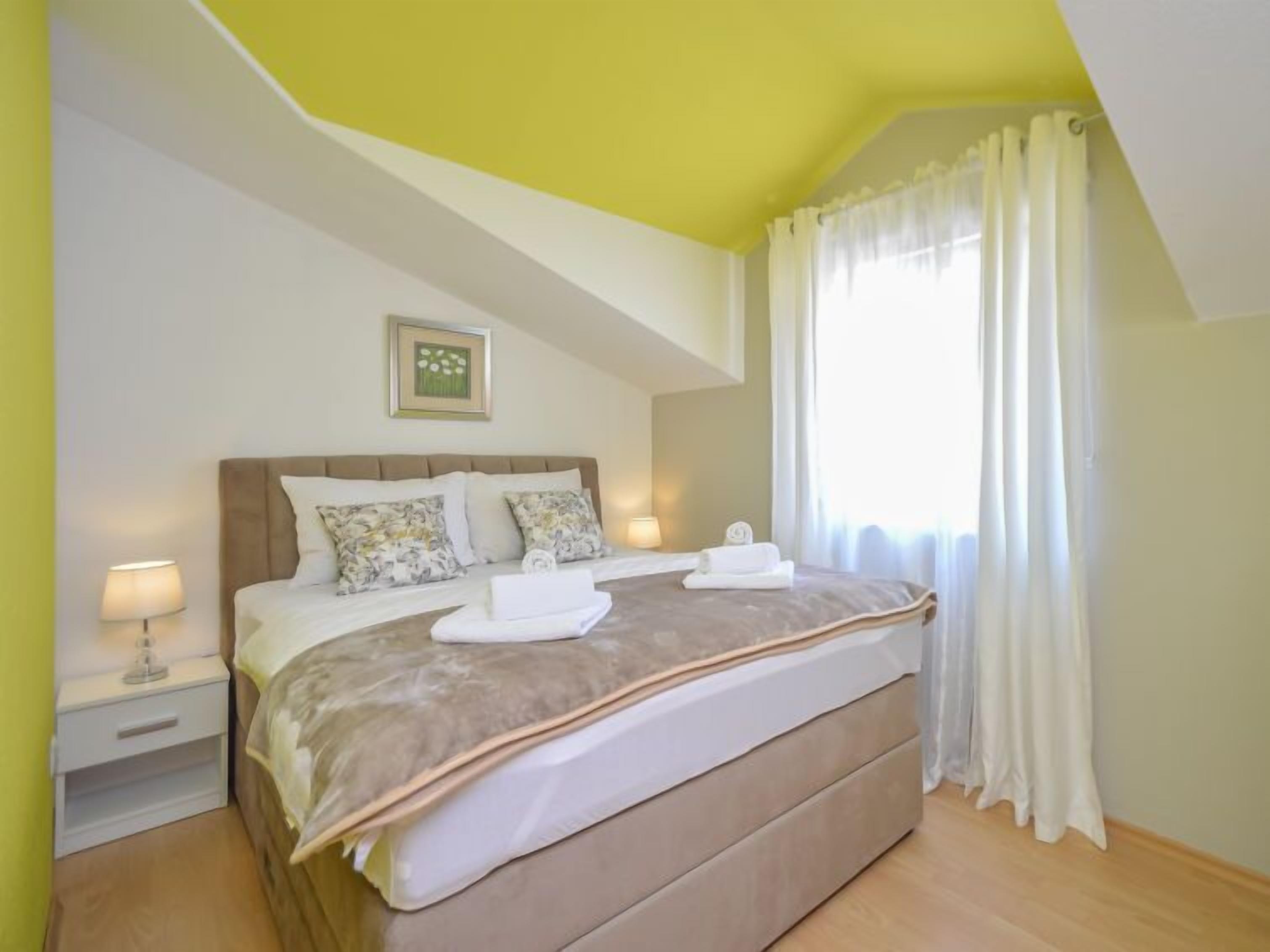 Een slaapkamer met een bed, nachtkastje, lamp, een ingelijst schilderij aan de muur en een raam met gordijnen.