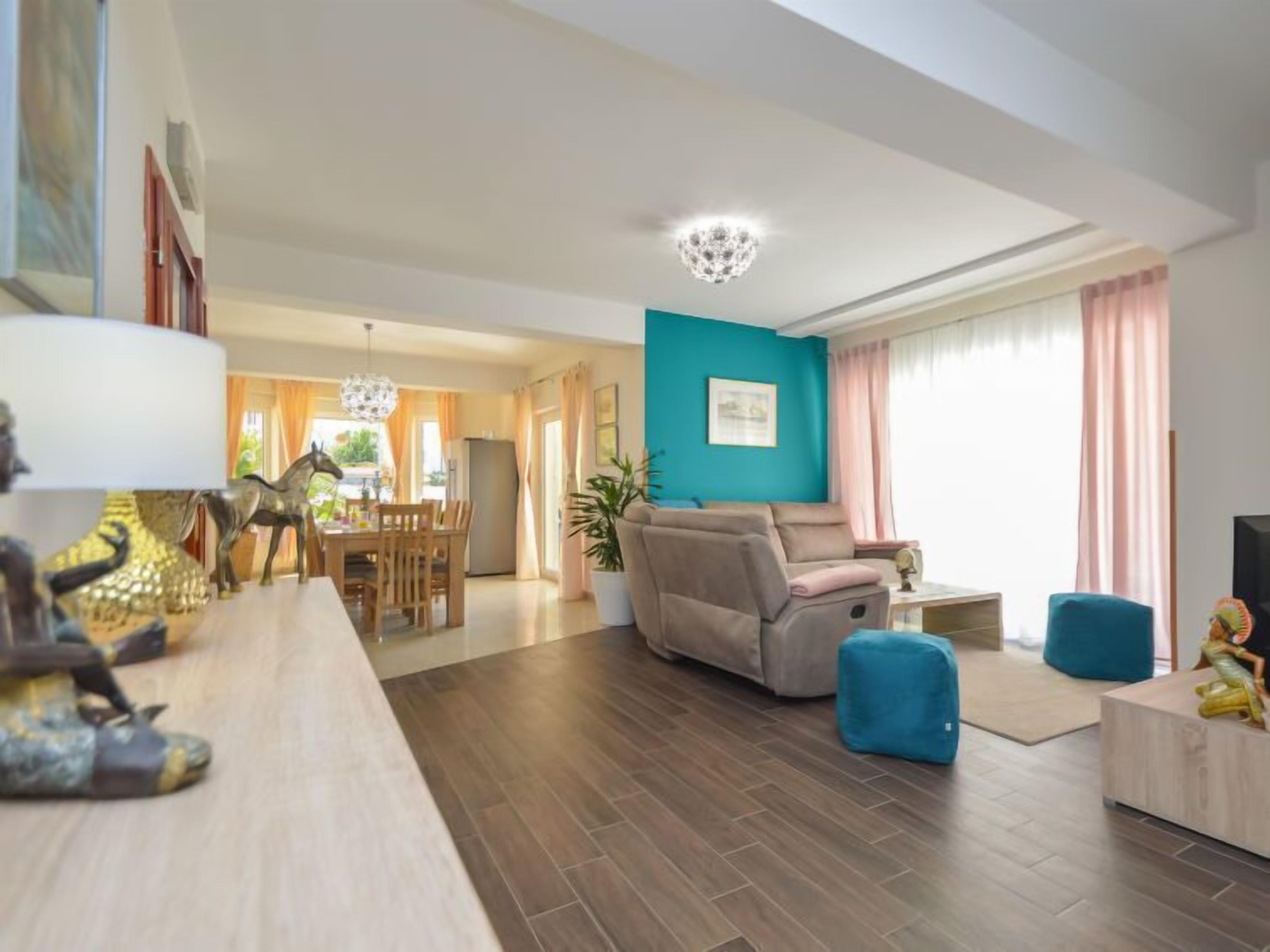 Een moderne woonkamer met een blauwgroene accentmuur, een beige bank, een eetgedeelte met houten meubels en een grote kroonluchter.