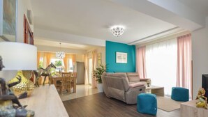 Ein modernes Wohnzimmer mit einer türkisfarbenen Akzentwand, einem beigen Sofa und einem Essbereich mit Holzmöbeln sowie einem großen Kronleuchter.