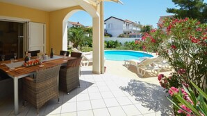 Eine überdachte Terrasse mit Esstisch, Korbstühlen und einem Pool im Hintergrund.