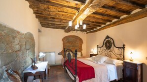 Une chambre avec un mur en pierre, un plafond en bois, une cheminée en briques, un lit à baldaquin, une table de chevet, une chaise et un bureau avec une lampe.