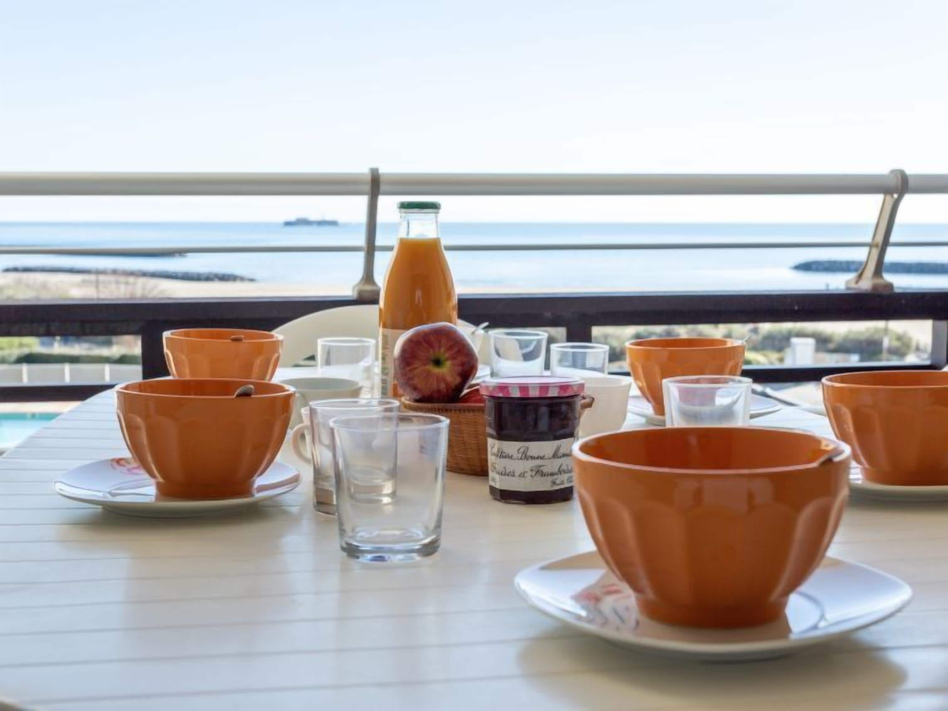 Ein Tisch ist gedeckt mit orangenen Bechern, einem Glas und einer Saftflasche, mit Blick auf ein Gewässer.