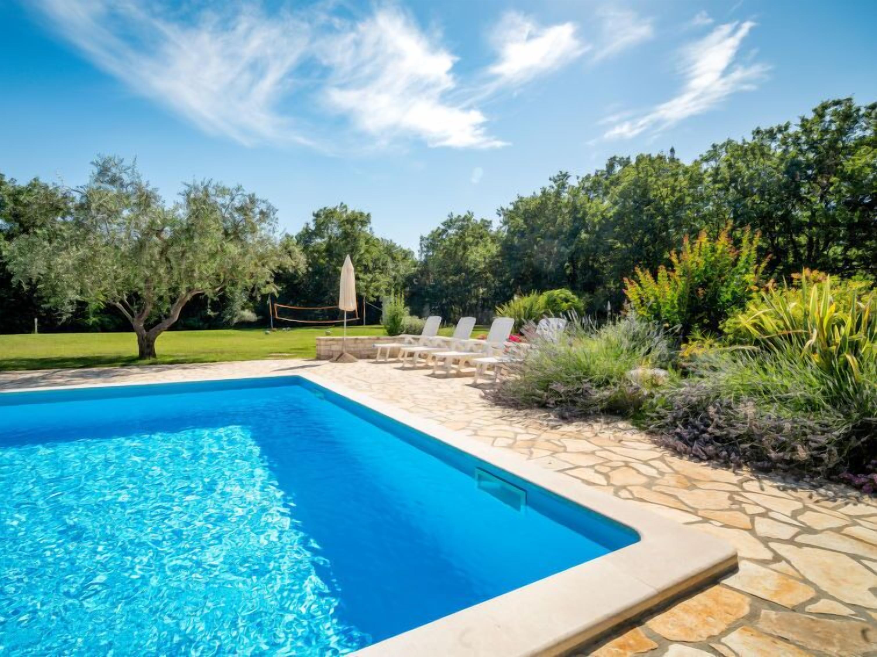 Une piscine rectangulaire à l’eau d’un bleu limpide, entourée d’une terrasse en pierre avec des chaises longues et d’une pelouse agrémentée d’arbres.