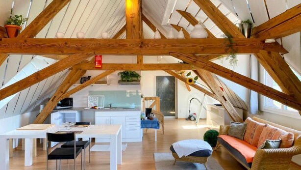 Un loft spacieux avec un plafond haut, des poutres apparentes, un coin cuisine, une table à manger avec des chaises et un coin salon confortable avec des coussins.