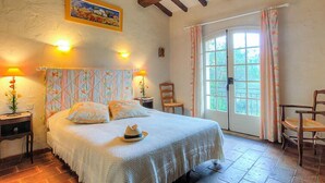Un dormitorio acogedor con cama, dos lámparas de noche, una silla de madera, una mesita pequeña y un gran ventanal con cortinas.
