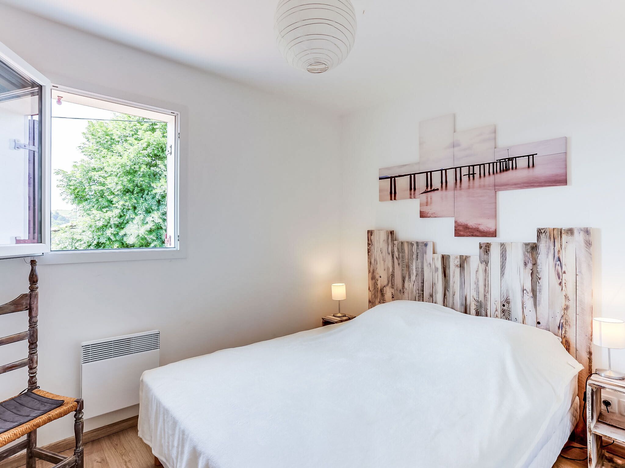 Ein Schlafzimmer mit einem Bett, einem Holzstuhl, einem Nachttisch mit Lampe, einem Heizkörper und einem Fenster mit Blick auf Bäume.