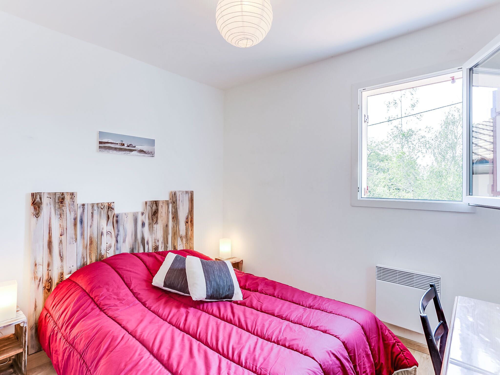 A bedroom with a wooden headboard, a red bedspread, a bedside table with a lamp, and a window with a view of trees.