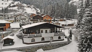 Un pueblo en la montaña cubierto de nieve, con edificios de estilo chalet, un muro de piedra y una valla de madera.