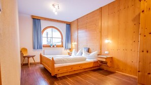 Una habitación con paneles de madera, una cama, una silla, una mesita y un ventanal con cortinas.