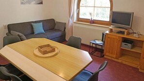 Una habitación acogedora con una mesa de comedor de madera, un sofá gris con cojines, un televisor sobre un mueble de madera y una ventana con cortinas.