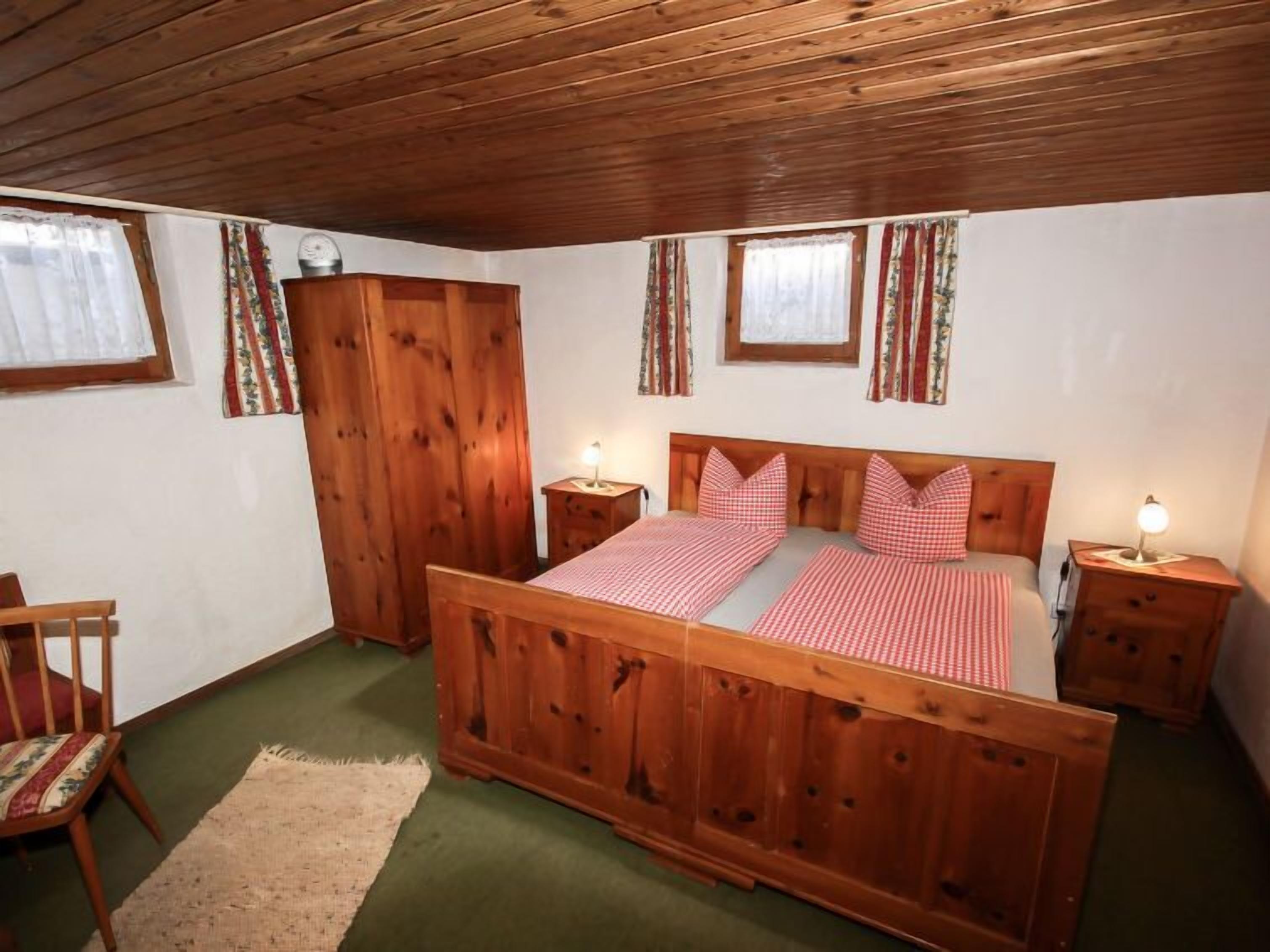Una cama con dosel de madera, con un edredón a cuadros, dos mesitas de noche con lámparas, un armario de madera y una silla con cojín estampado.