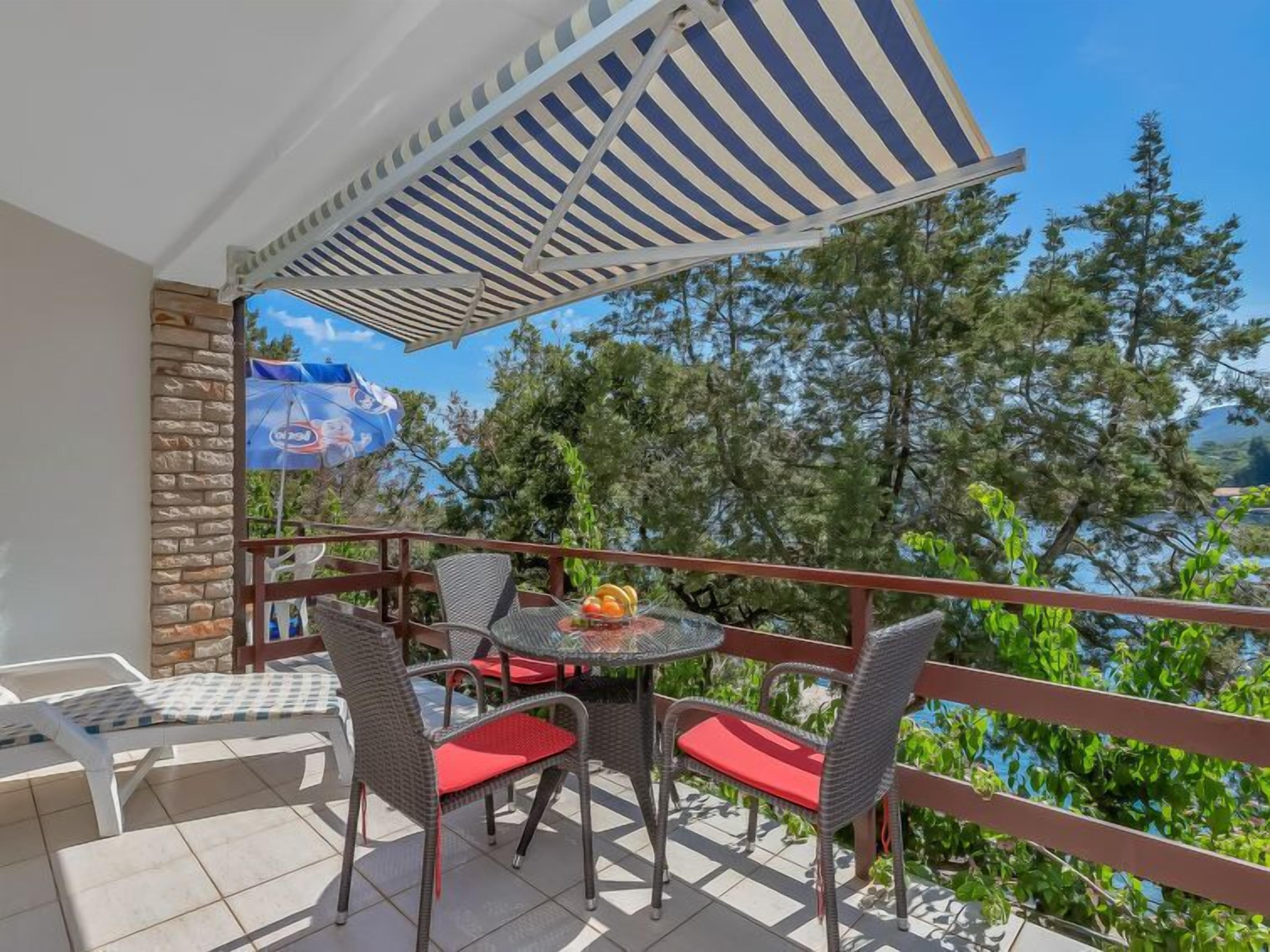 A balcony with wicker chairs and a table, a striped awning, and a view of trees and a body of water.