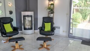 Deux fauteuils noirs avec des coussins verts, une cheminée à foyer vitré et une vue sur la verdure à l’extérieur.