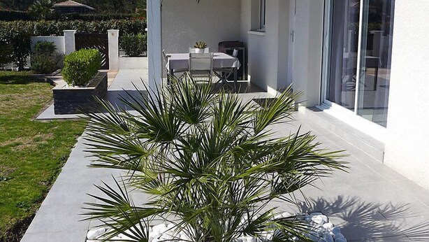 Une maison moderne dotée d’un patio comprenant une table et des chaises, entouré de plantes en pot et d’une pelouse bien entretenue.