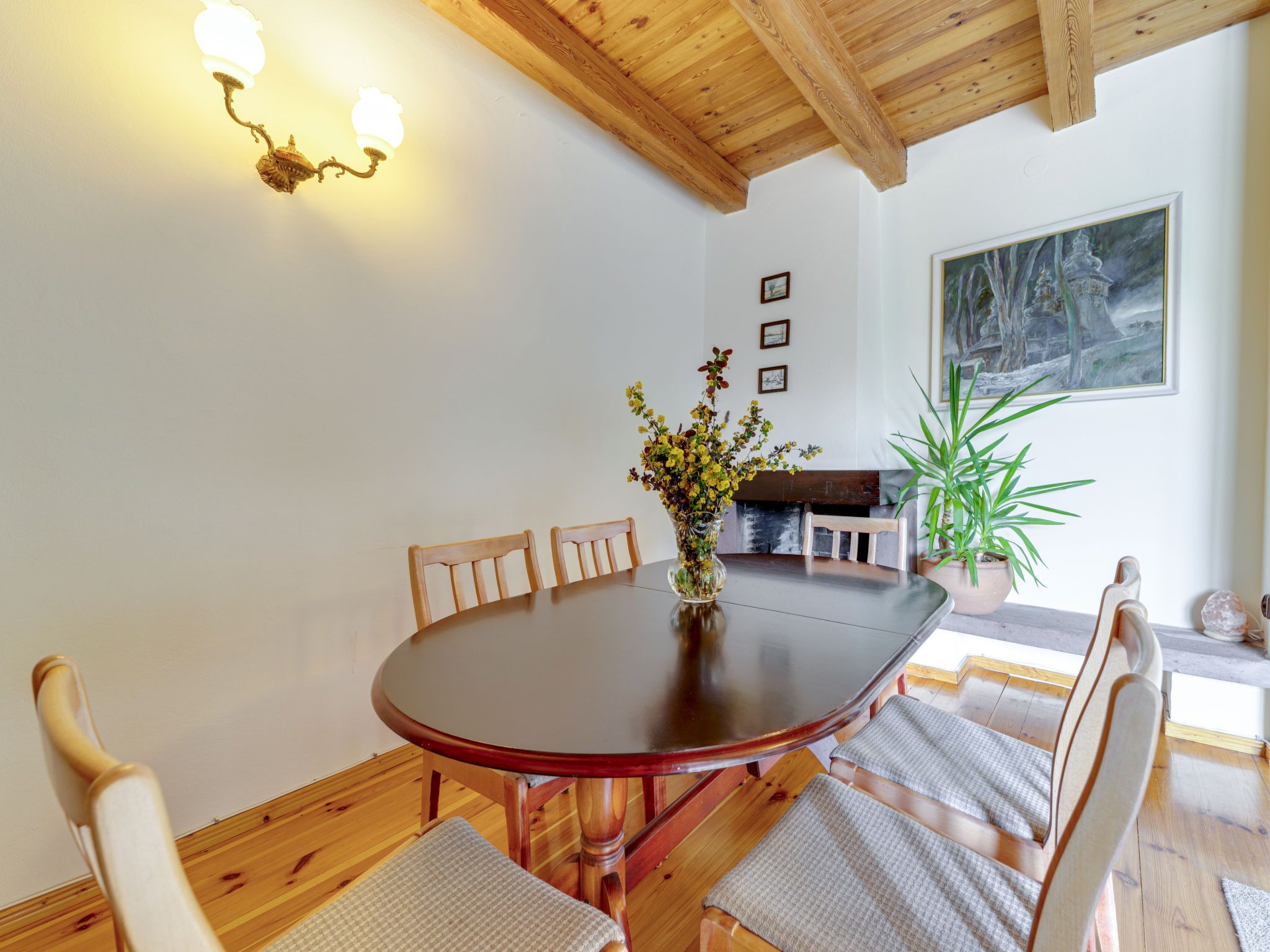 A dining area with a wooden table, chairs, a vase with flowers, and a painting on the wall.