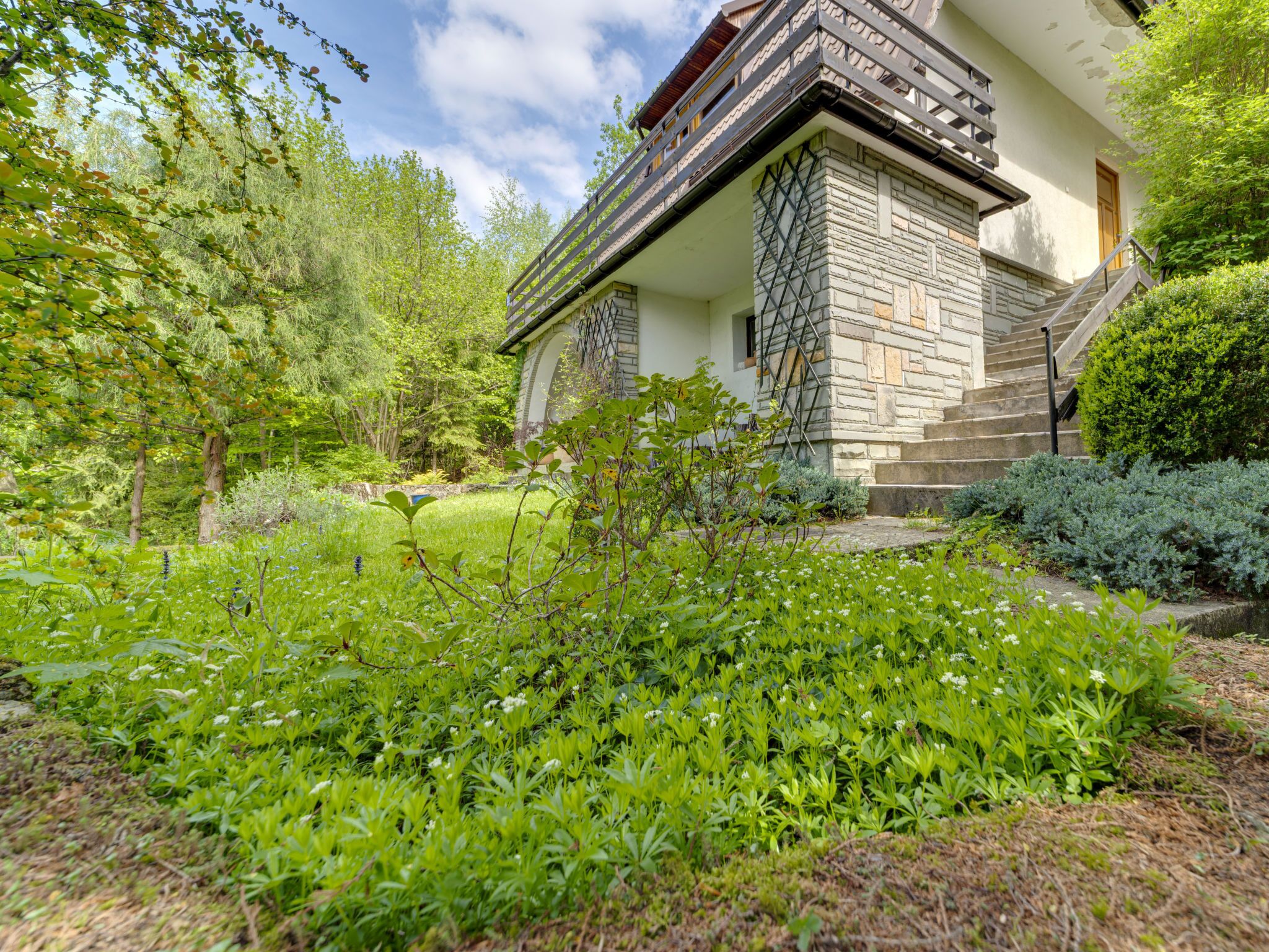 A house with a stone facade, a wooden balcony, and a well-maintained garden with various plants and a lawn.
