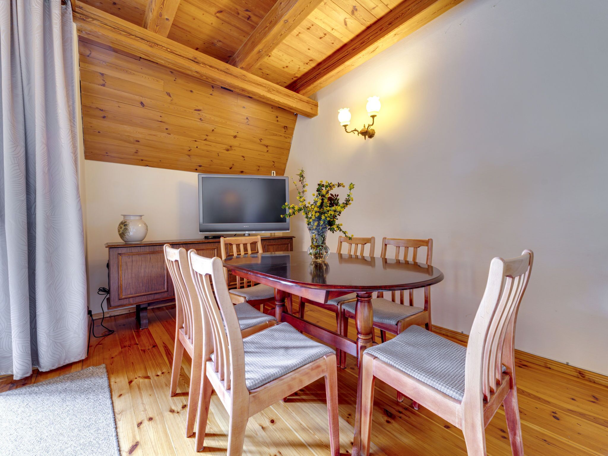 Un coin repas avec une table en bois, des chaises, un téléviseur et un vase rempli de fleurs.