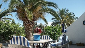 Une terrasse extérieure avec des chaises à rayures et une table, entourée de palmiers et d’un mur blanc.
