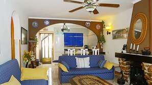 Un salon avec un canapé bleu, une table basse en bois et une cheminée. Au fond, on aperçoit une cuisine avec des meubles bleus et un coin repas.