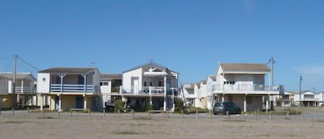 A row of beach houses with balconies, a sandy foreground, and a clear blue sky.