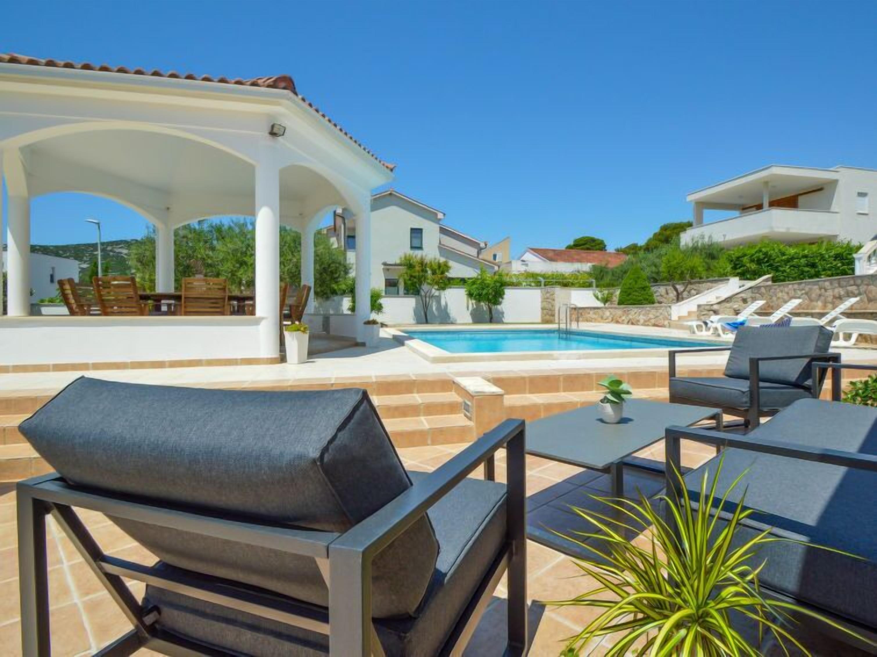Ein Poolbereich mit Außenmöbeln, einer überdachten Terrasse und Blick auf den Pool sowie die umliegenden Gebäude.