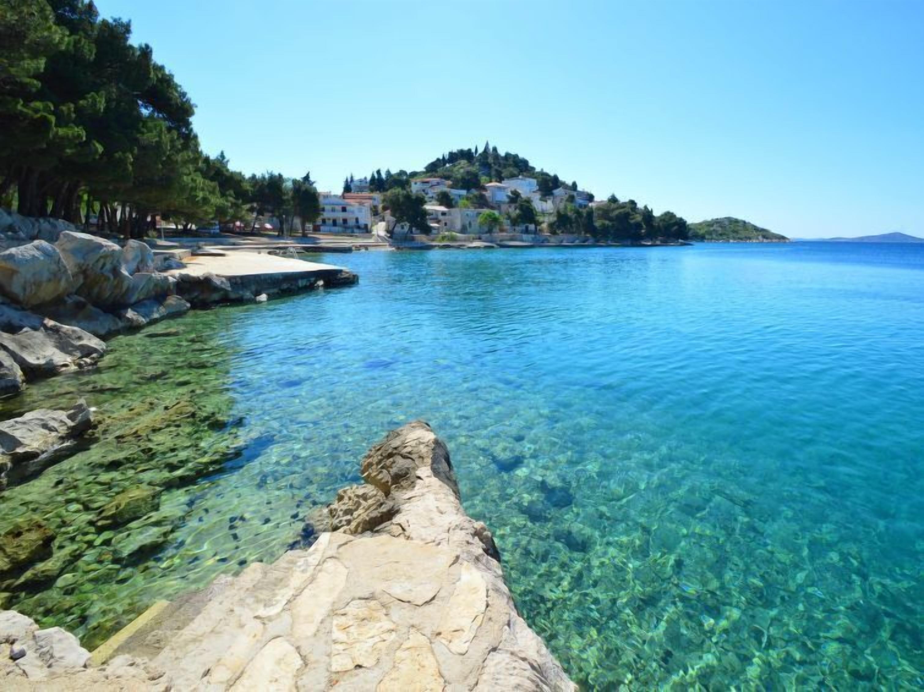 A clear coastal view with rocky shoreline, clear water, and a hill with buildings in the background.