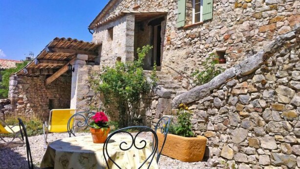Un bâtiment en pierre à l’aspect rustique, doté d’un petit balcon et d’un espace extérieur aménagé avec des chaises et une table jaunes.