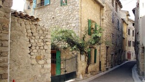 Une rue étroite bordée de bâtiments en pierre, avec un arbre au milieu, une clôture en métal et une porte verte.