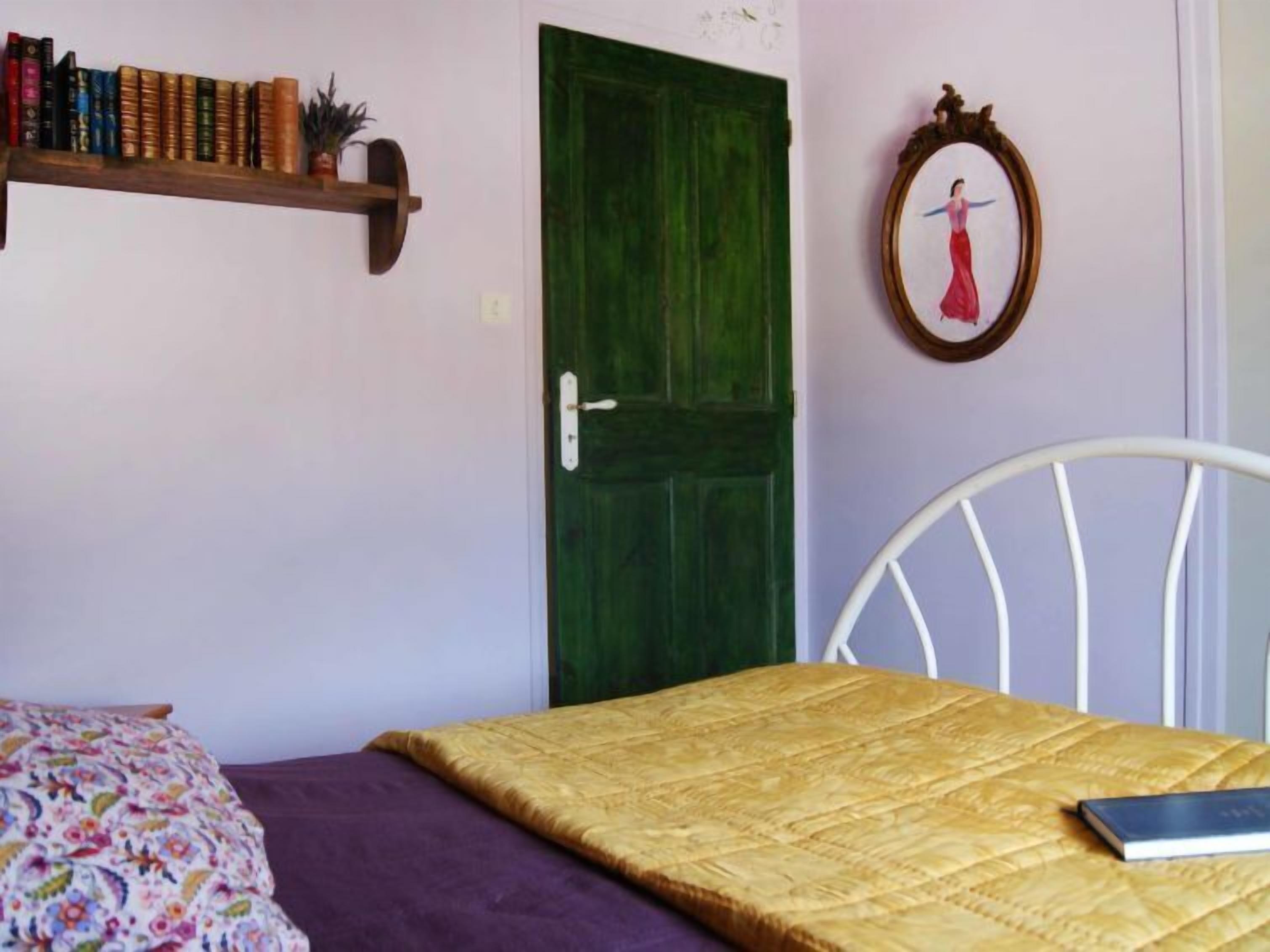 A bedroom with a bed, a chair, a bookshelf, a green door, and a framed picture on the wall.