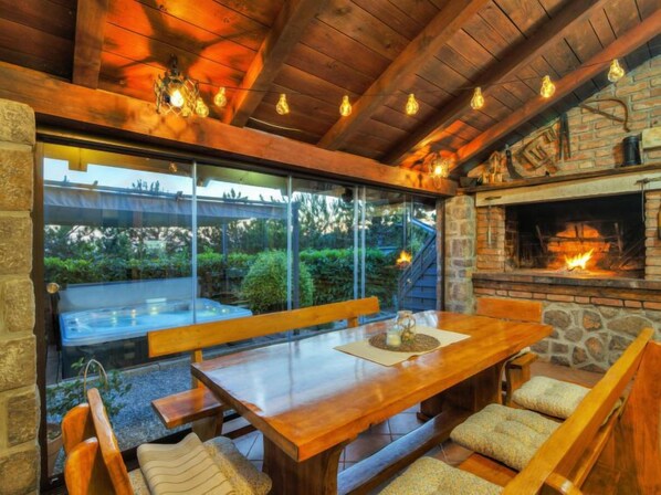 An outdoor dining area with a wooden table and benches, a stone fireplace, and a glass enclosure with a view of greenery. - Rukavina by Interhome (Senj)