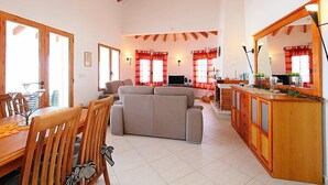 Een ruime woonkamer met een bank, eettafel en stoelen. Er is een keukenhoek met een aanrecht en kasten.