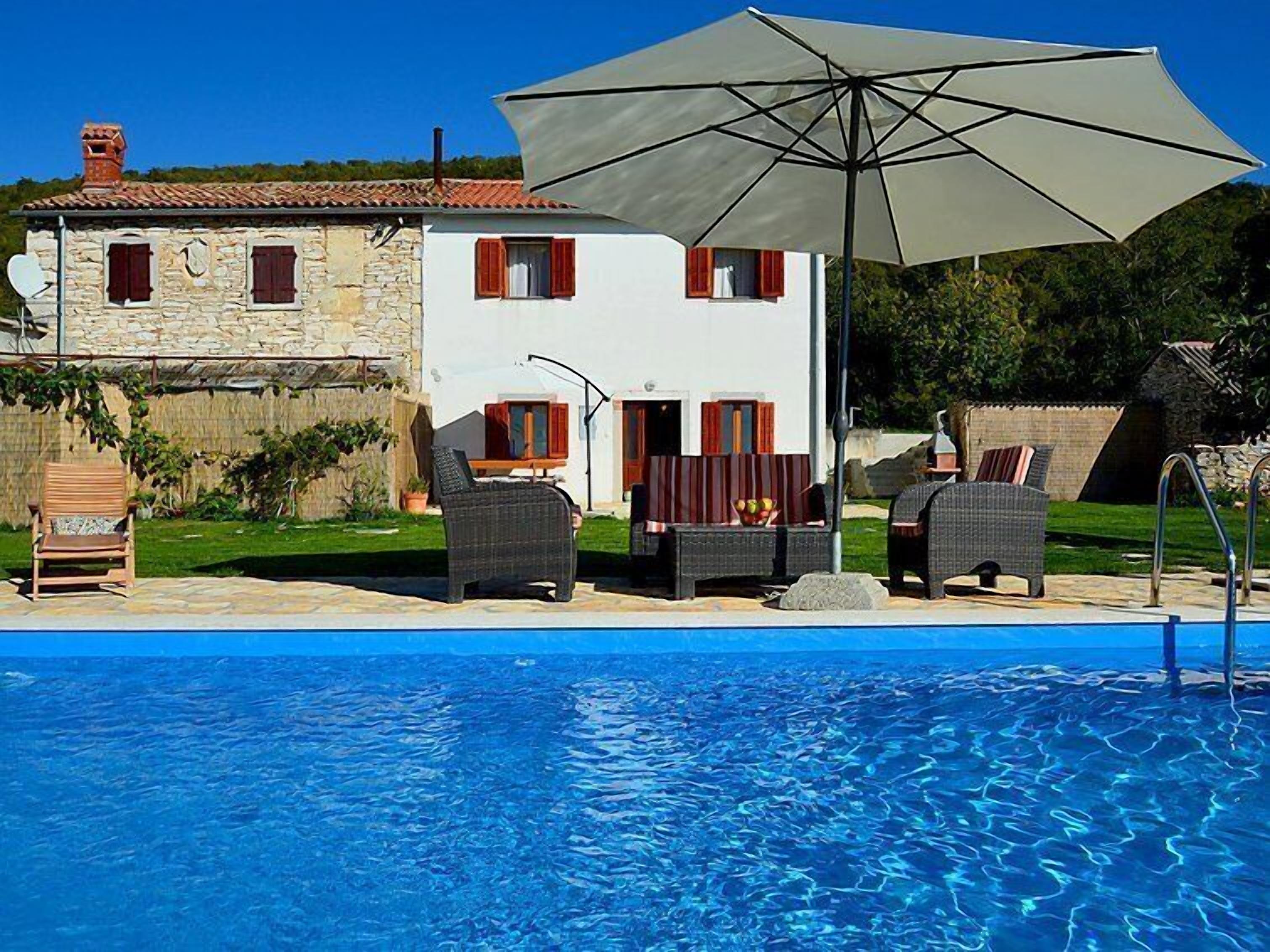 A poolside area with a stone house, outdoor furniture, and a large umbrella.