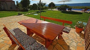 Eine Terrasse im Freien mit einem Holztisch und Bänken, einem großen Sonnenschirm und Blick auf ein Schwimmbad und Berge im Hintergrund.