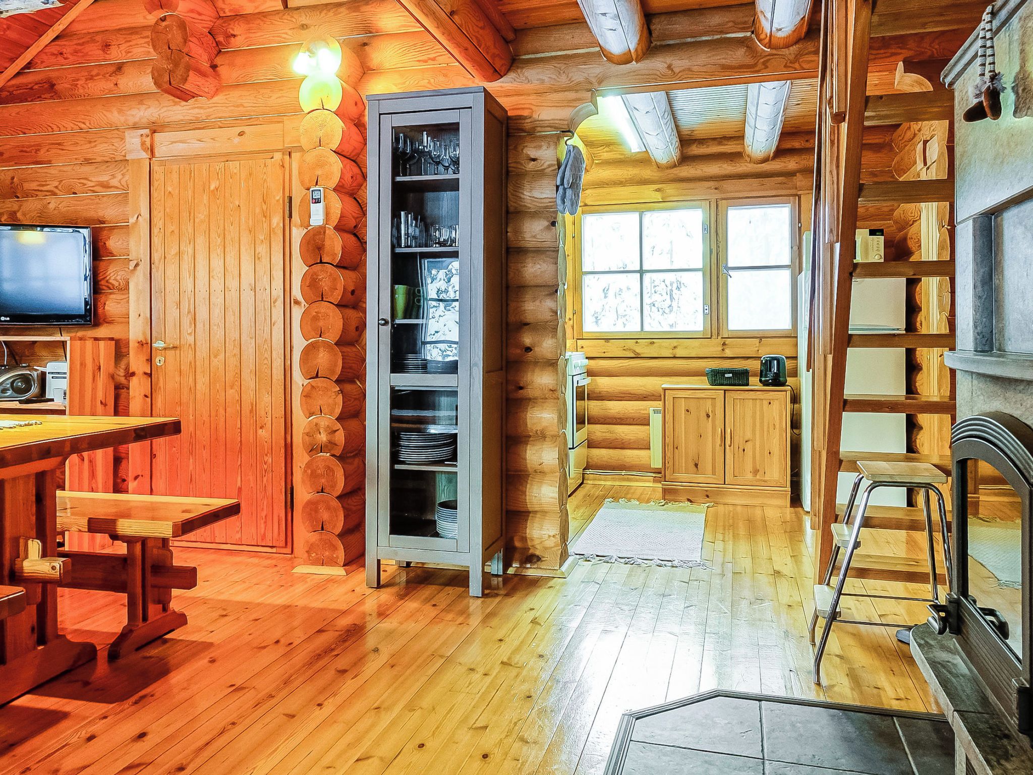 A log cabin interior with a wooden dining table, a built-in bookshelf, a fireplace, and a window with a view.