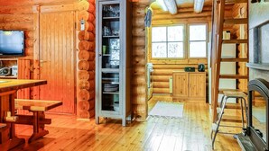 A cozy log cabin interior with a wooden dining table, a built-in cabinet with shelves, a kitchen area with a stove and sink, and a television on the wall.