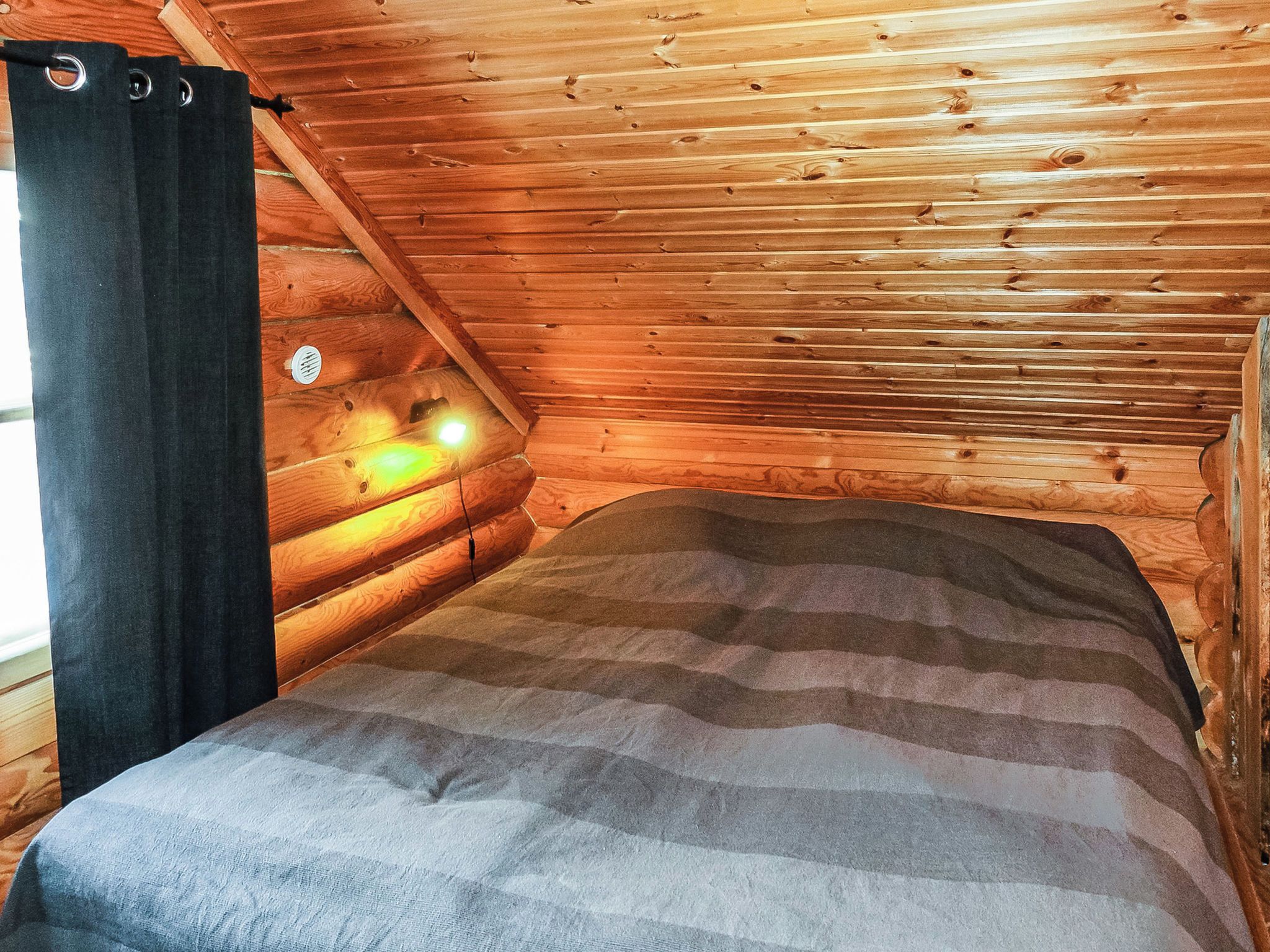 Puuinen mökki, jossa on sänky, ikkuna verhoilla ja seinään kiinnitetty valaisin.